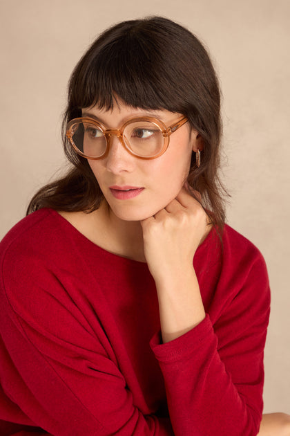Woman with brown hair wearing Crystal Champagne Round Reading Glasses and a red top, resting her chin on her hand, looking to the side against a plain background.