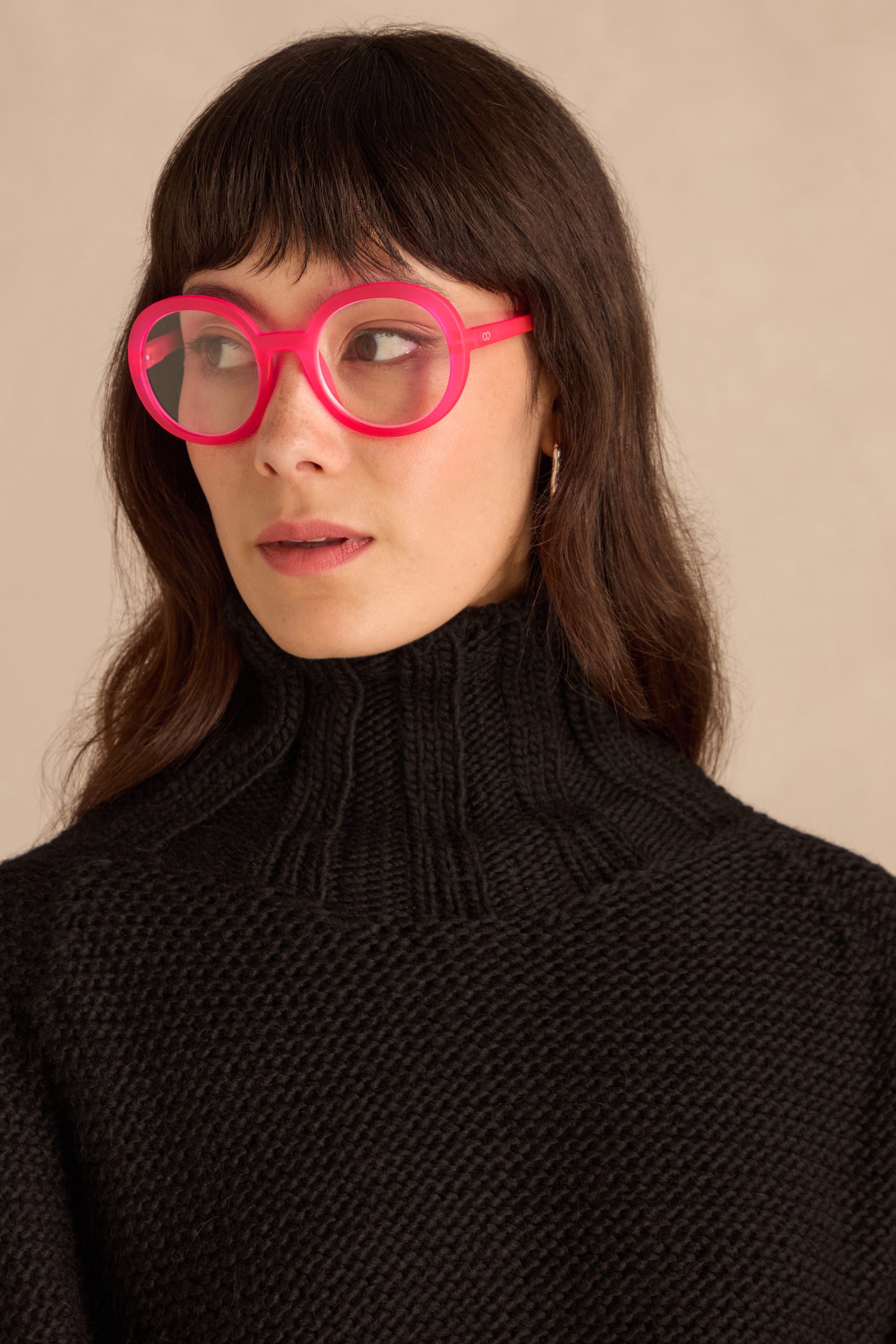 Woman with long brown hair in a black turtleneck wears Neon Pink Round Reading Glasses with optician-quality lenses, looking to the side against a neutral background.