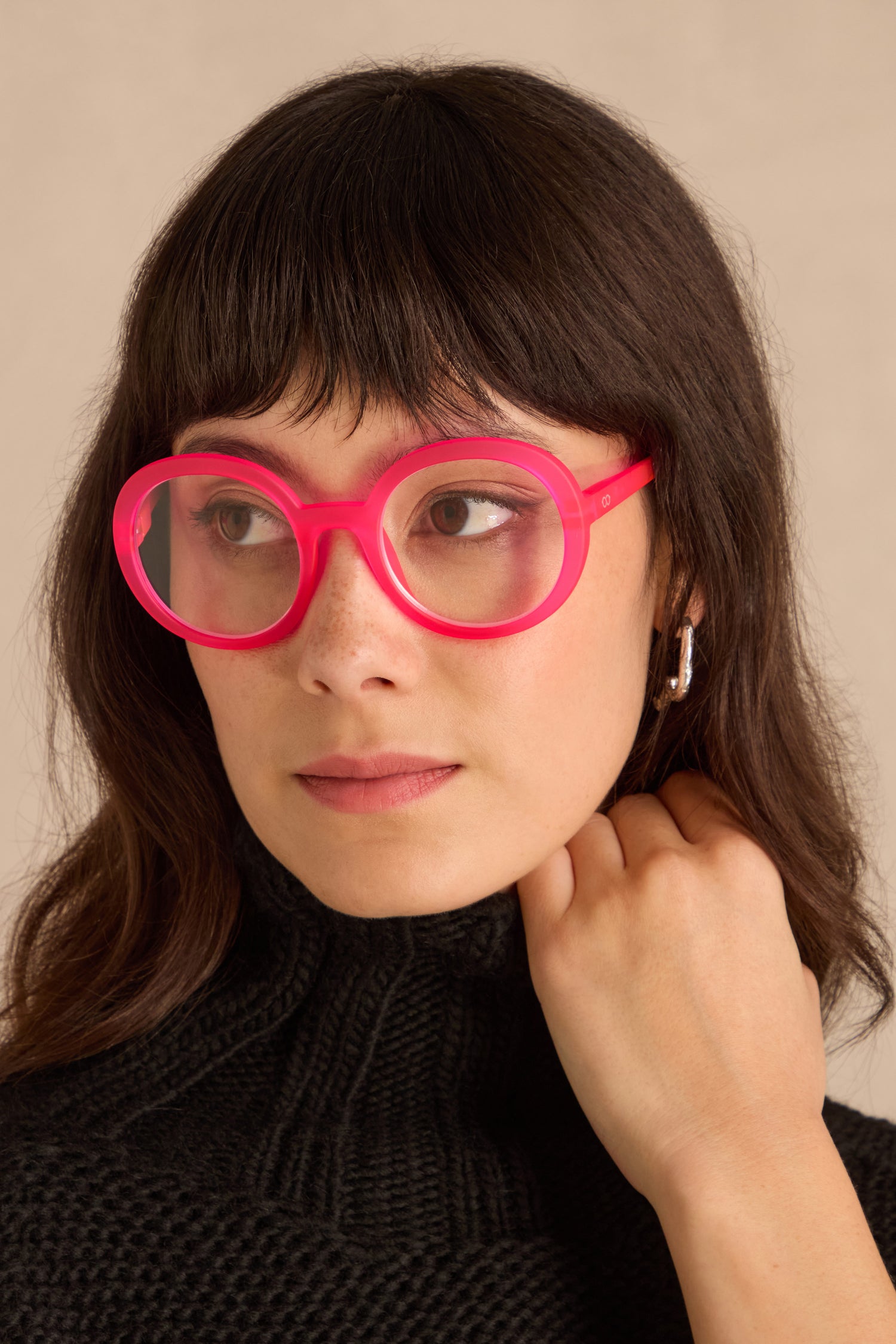 Woman with shoulder-length brown hair wears Neon Pink Round Reading Glasses and a black turtleneck, looking to the side with her hand on her neck.