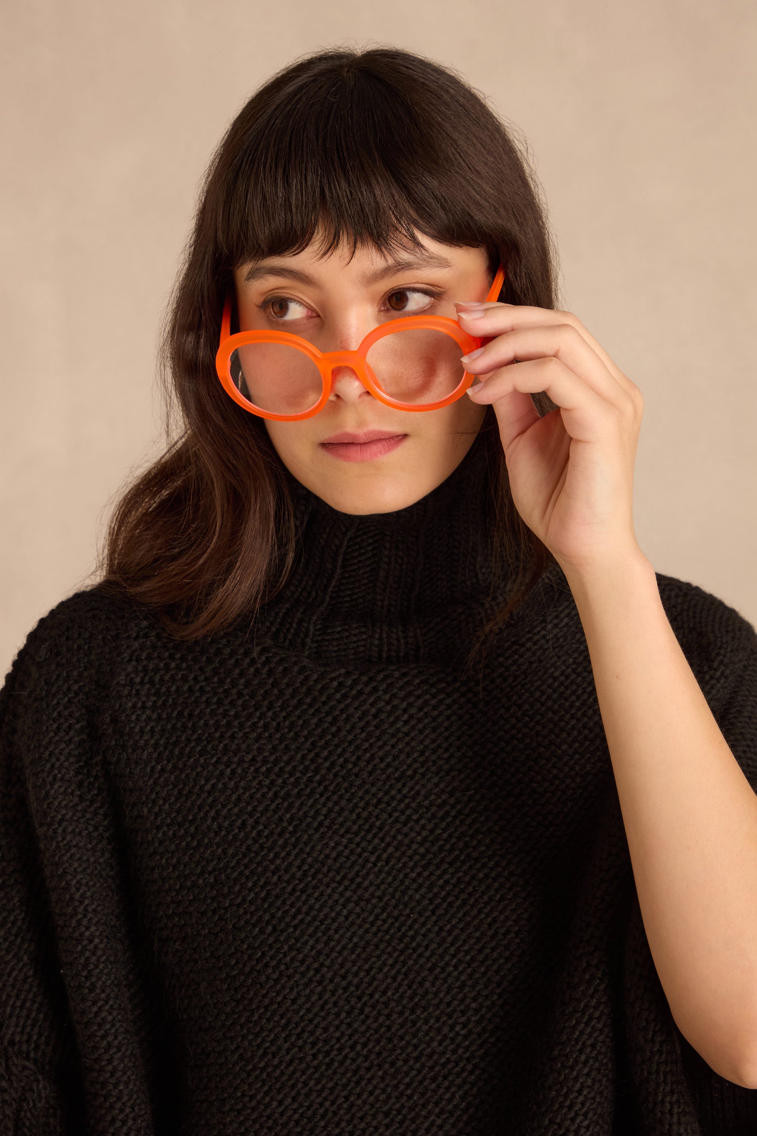 A person with long dark hair and bangs wears a black sweater and Neon Peach Round Reading Glasses, looking to the side while adjusting the optician-quality lenses.