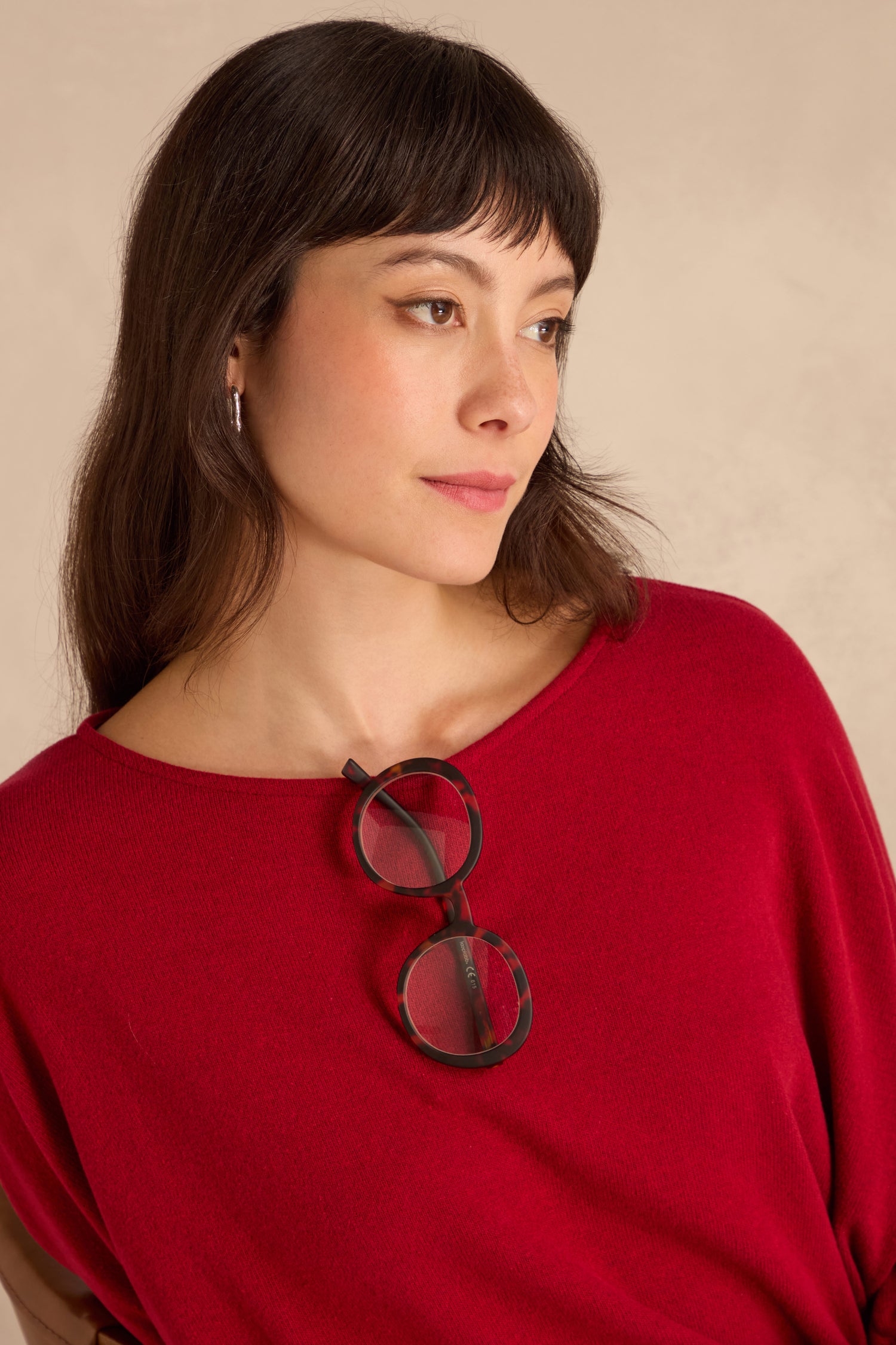 Woman with straight brown hair in a red top and silver hoop earrings, looking to the side with Tortoise Round Reading Glasses hanging from her neckline.