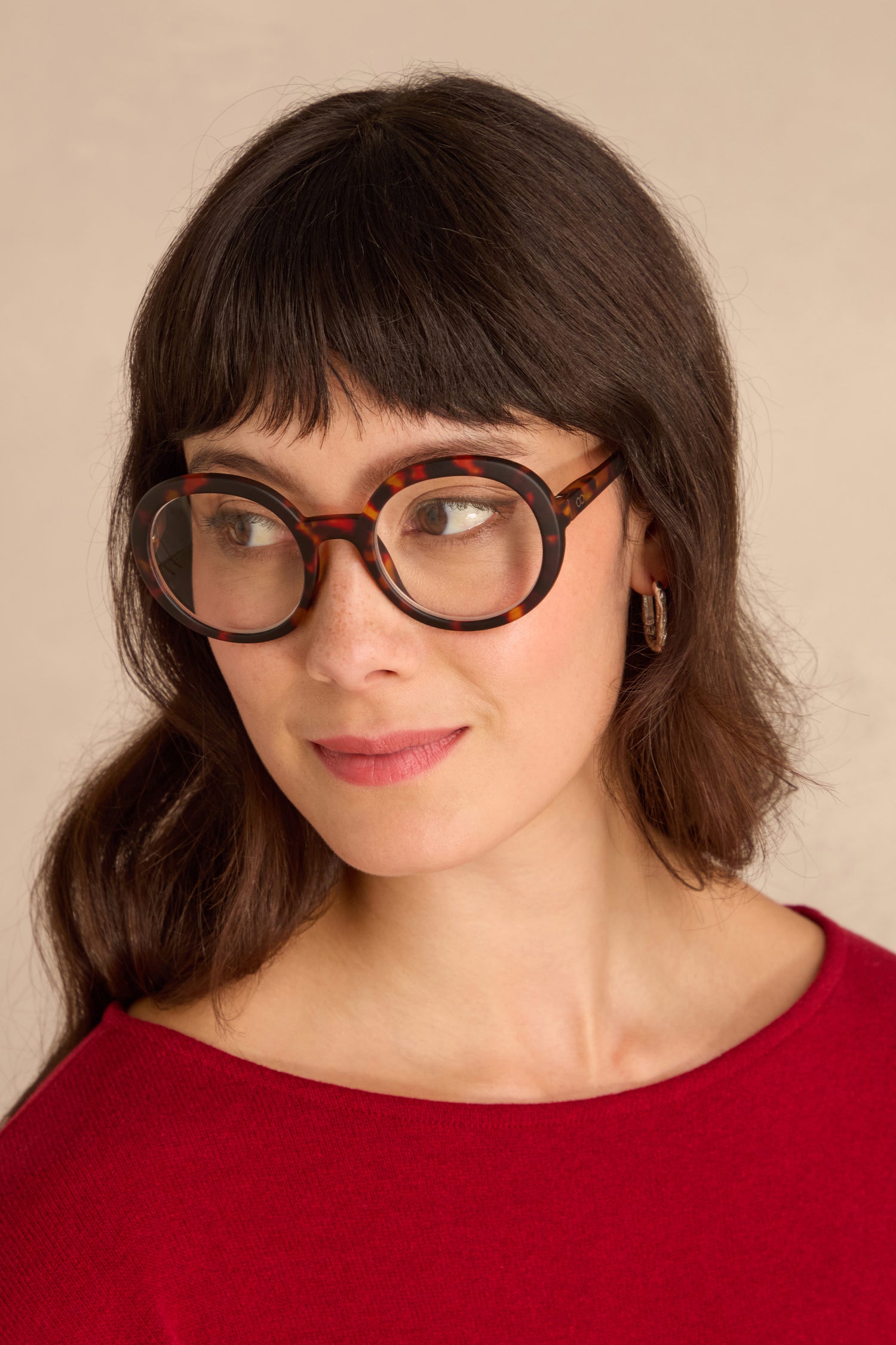 Woman with long brown hair and a red top wears Tortoise Round Reading Glasses, looking slightly to the side against a neutral background.