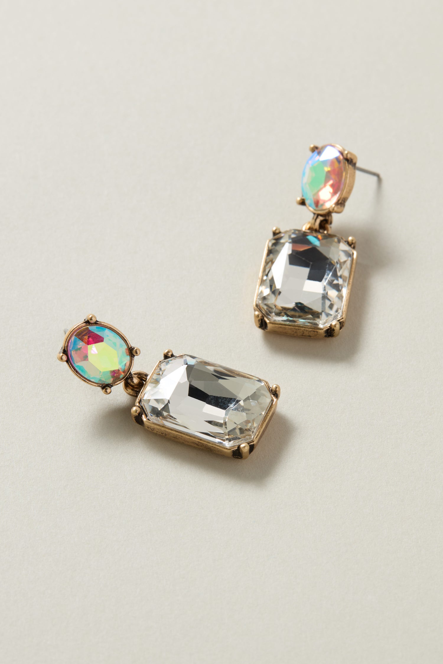 The Twin Gemstone Earrings are luxurious accessories featuring large rectangular clear crystals and smaller iridescent oval stones, creating a stunning contrast of vibrant color against a light background.