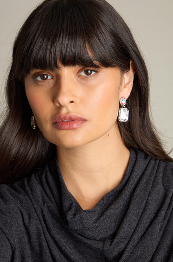 A person with long dark hair and bangs, wearing a dark top and the Crystal Gemstone Earrings featuring iridescent oval stones, stares ahead.