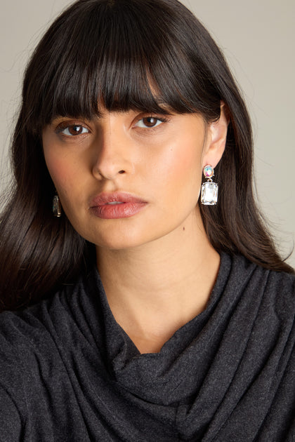 A person with long dark hair and bangs, wearing a dark top and the Crystal Gemstone Earrings featuring iridescent oval stones, stares ahead.