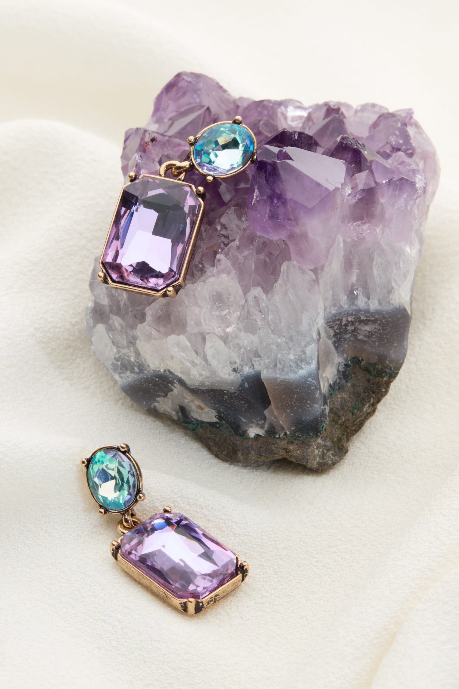 The Twin Gemstone Earrings, featuring large rectangular lilac stones and smaller round azure stones, are beautifully displayed on a piece of amethyst crystal against a soft white fabric backdrop.