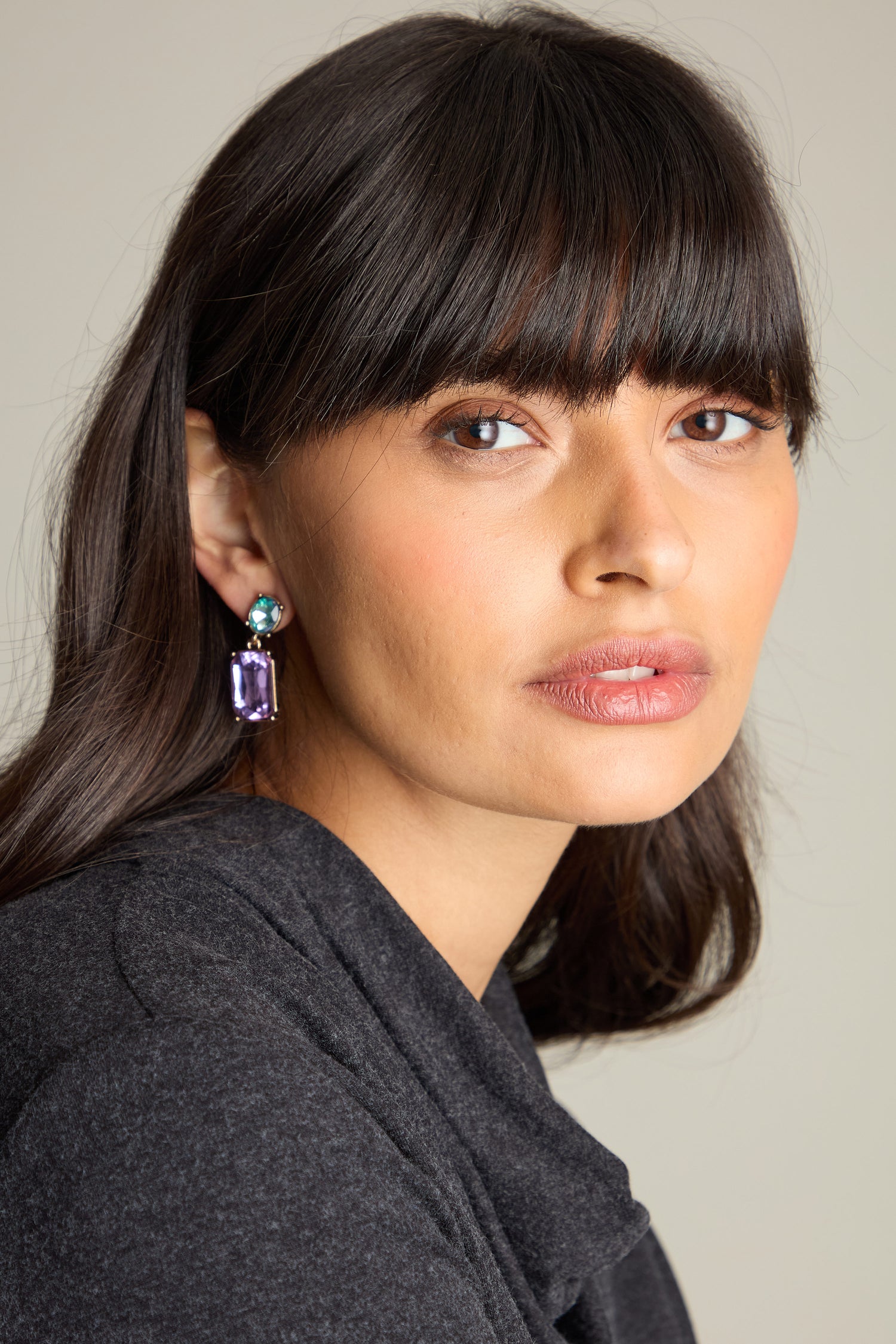 A person with shoulder-length brown hair and bangs wears a grey top, showcasing the luxurious Twin Gemstone Earrings adorned with lilac and azure stones, looking slightly to the side.