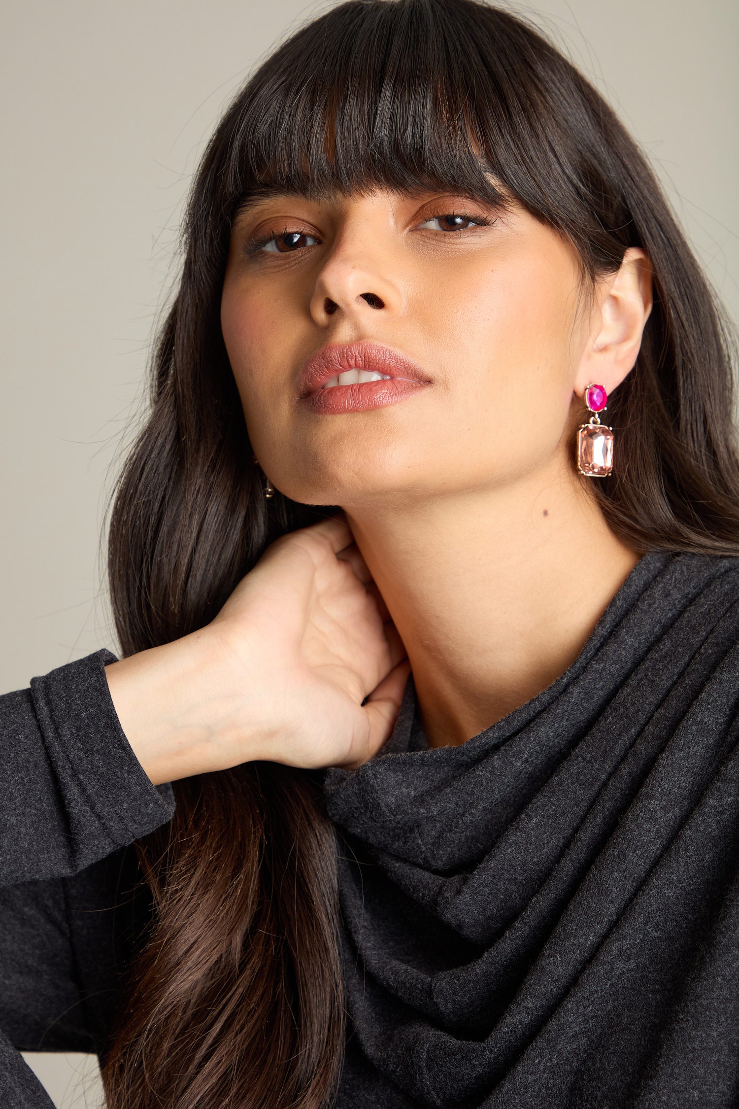 A woman with long, dark hair and bangs, wearing a dark gray top and Twin Gemstone Earrings, rests one hand on her neck while looking directly at the camera.