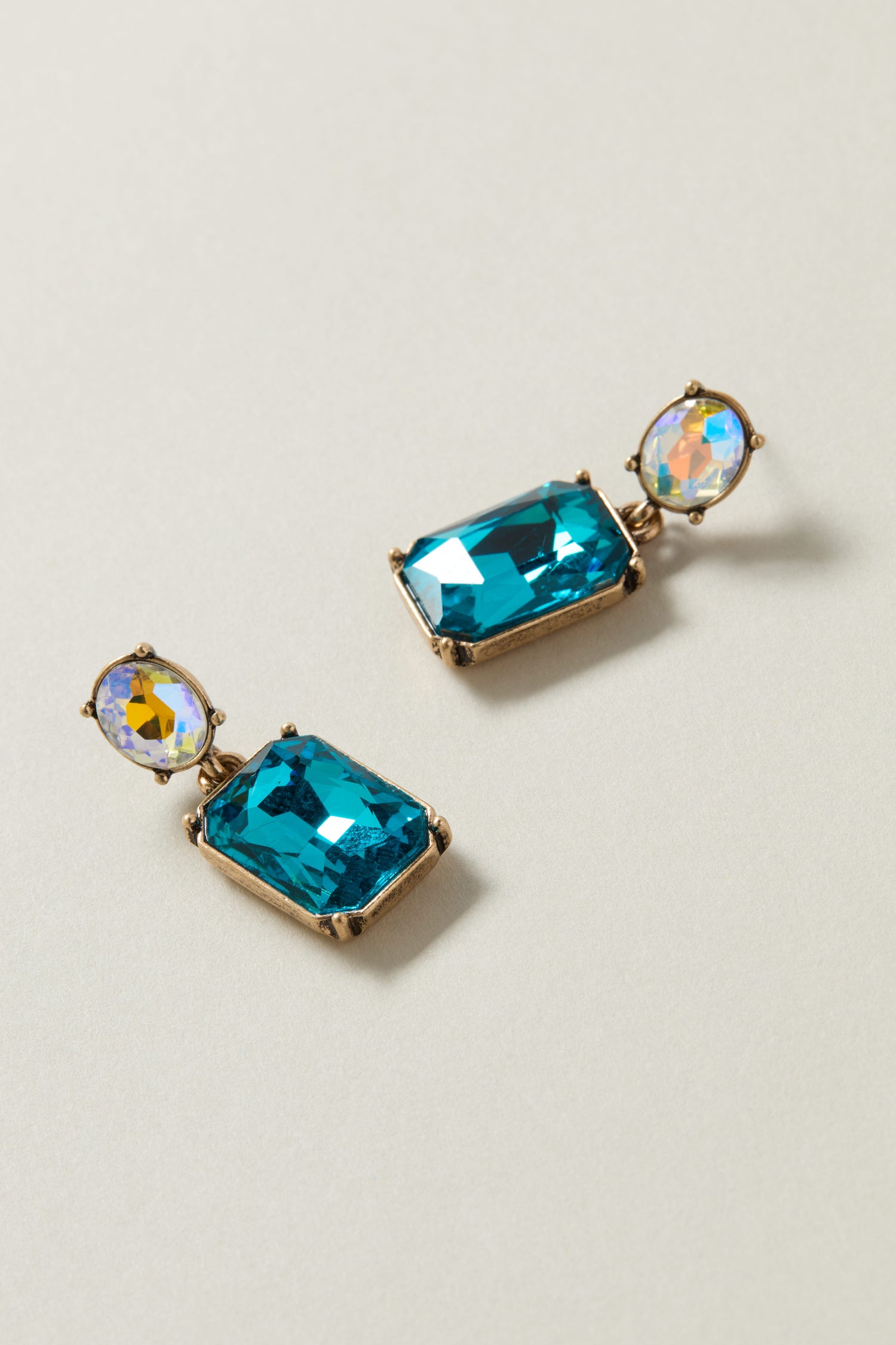 The Twin Gemstone Earrings each showcase a petite, round, iridescent gemstone nestled at the top and a larger, rectangular azure gemstone suspended below, all framed in luxurious gold. These exquisite pieces of jewelry seamlessly combine elegance with vivid colors against a light backdrop.