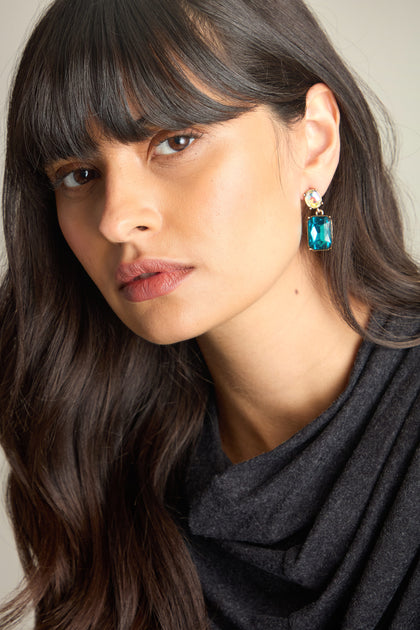 A person with long, dark hair and bangs is wearing a black top and luxury jewelry, featuring Twin Gemstone Earrings adorned with lilac and azure gemstones, looking directly at the camera.