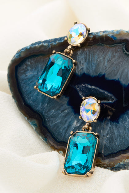 The Twin Gemstone Earrings, featuring a combination of large azure gemstone pendants and smaller iridescent stones, are elegantly displayed on a sliced geode placed on beige fabric.