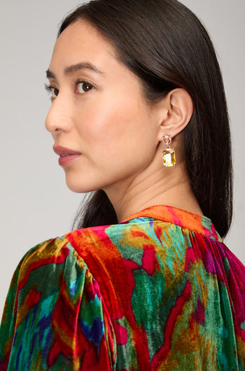 A woman with long dark hair wears striking Twin Gemstone Earrings featuring large rectangular yellow stones and a colorful velvet top, glancing over her shoulder against a plain background.