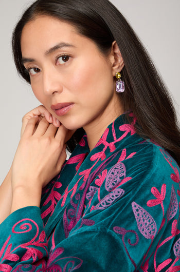 A woman with long dark hair wears Twin Gemstone Earrings and an embroidered teal and magenta garment, gazing calmly at the camera.