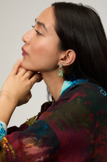 A woman in profile wears Twin Gemstone Earrings, featuring large green stones, paired with a colorful top as she rests her chin on her hand against a neutral background.