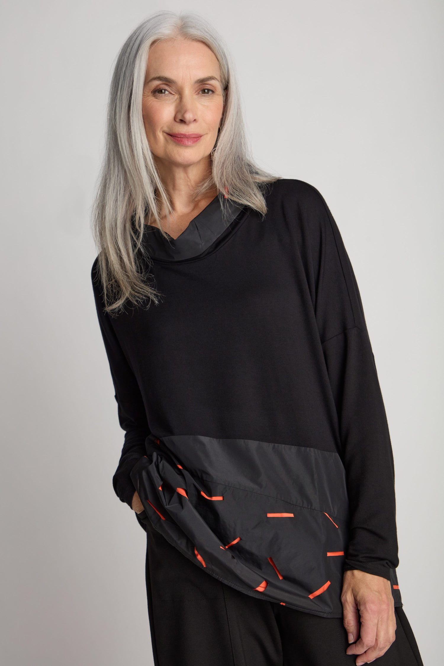 A woman with long gray hair is wearing a Cut Out Cowl Neck Tunic, featuring a black sweater with a patterned bottom section and showcasing a minimalist design. She is standing against a plain background.