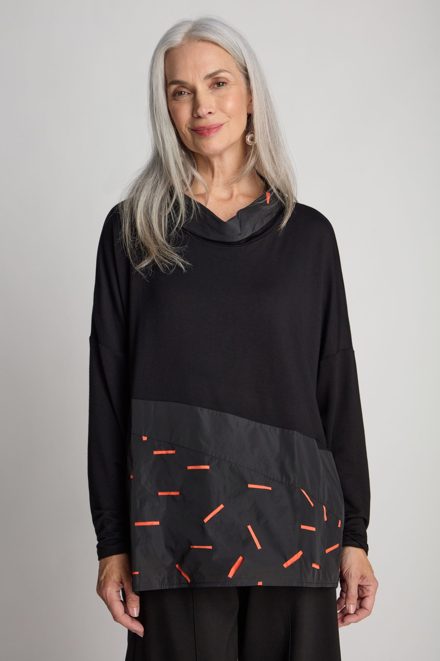 A woman with long gray hair and donning a black and red patterned Cut Out Cowl Neck Tunic stands against a plain background.