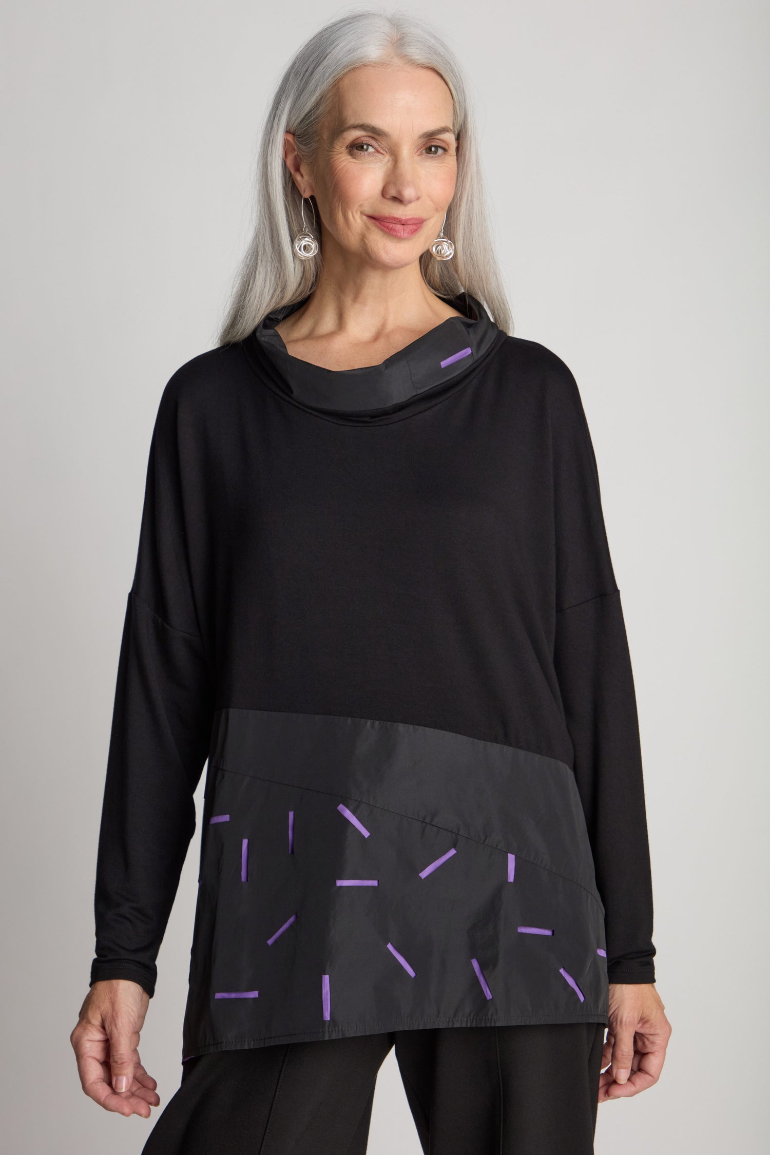 A woman with long gray hair stands against a plain background, wearing the Cut Out Cowl Neck Tunic—a black long-sleeved top with contrasting fabric adorned with purple accents. The minimalist design of the tunic complements her smile as she faces the camera.