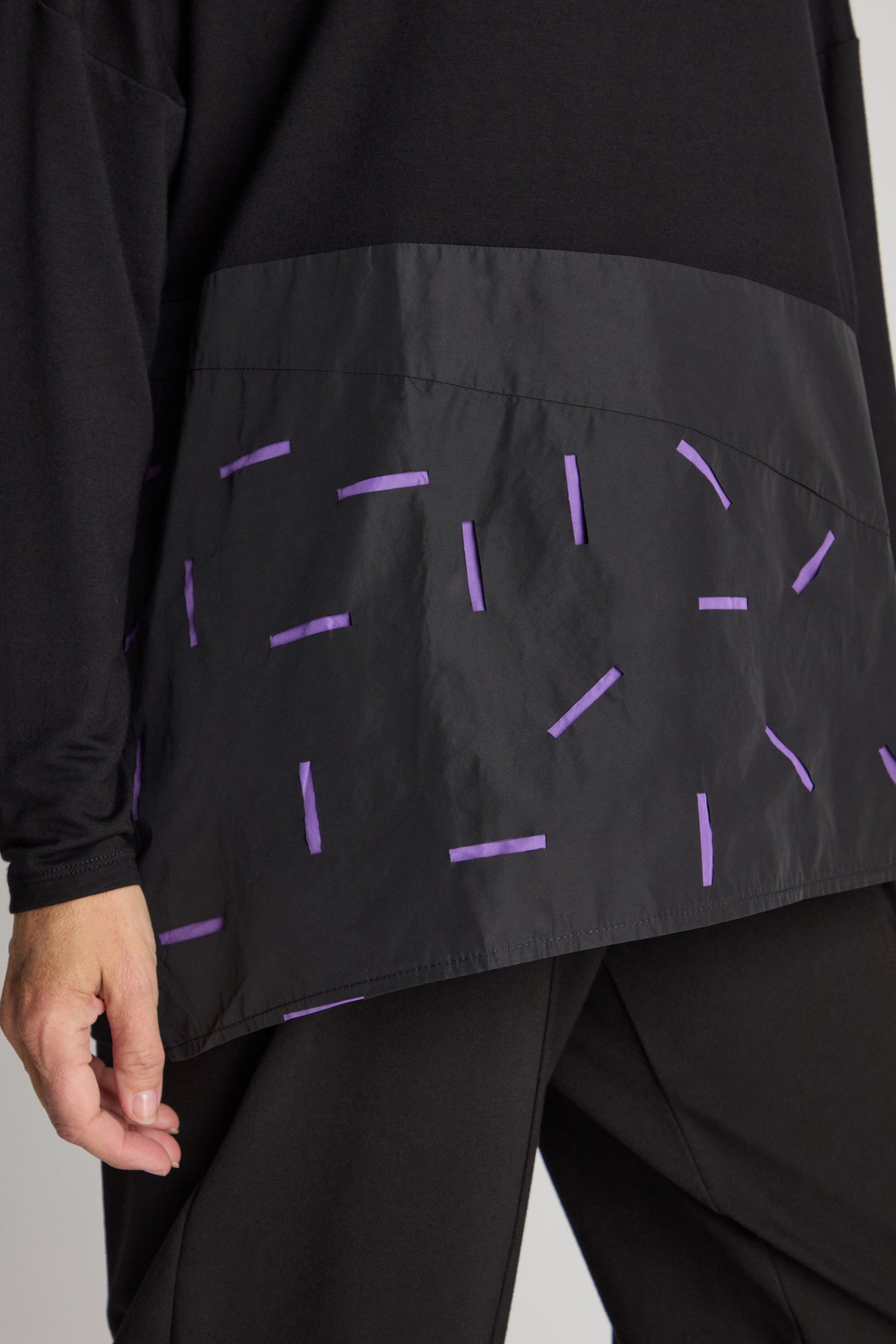 Close-up of a person wearing the Cut Out Cowl Neck Tunic, a black garment with a minimalist design featuring contrasting fabric adorned with scattered purple rectangular designs.