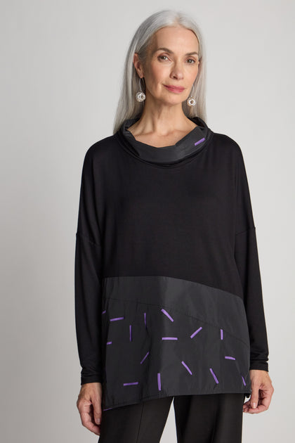 A woman with long gray hair wearing a cut out cowl neck tunic featuring black long-sleeve top with purple patterns stands against a plain, minimalist background, creating an elegant look.