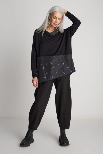 A person with long gray hair poses in the minimalist Cut Out Cowl Neck Tunic, paired with loose-fitting pants and black boots. The black outfit is accented with purple details, set against a plain background.