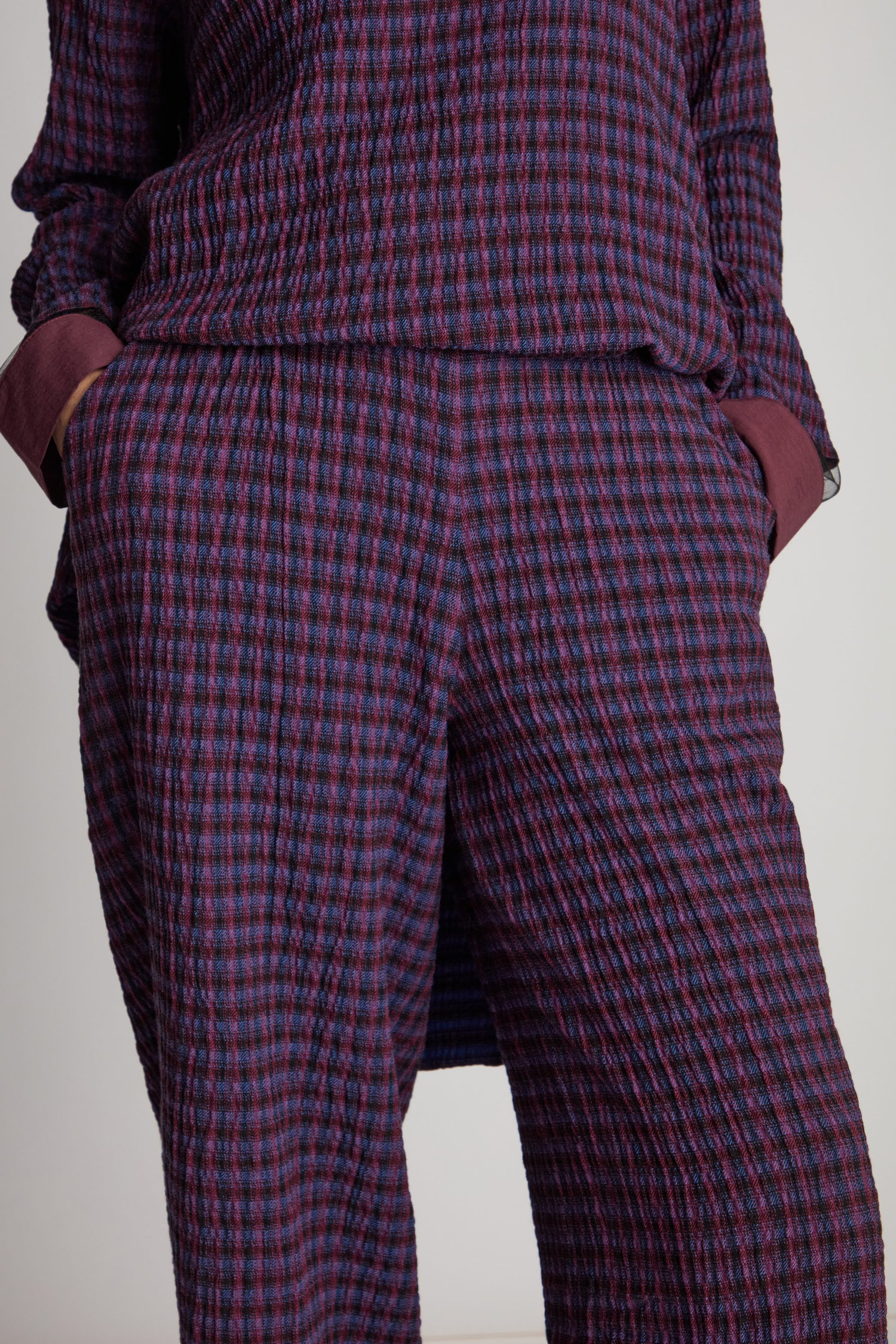 Person wearing Check Trousers, textured with a purple plaid pattern and made of gauze linen, showing the waist and hip area with hands in pockets.