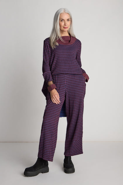 A woman with long gray hair wearing the Check Trouser paired with a matching purple and pink checkered top and black boots stands confidently against a plain background, embodying sustainable fashion.