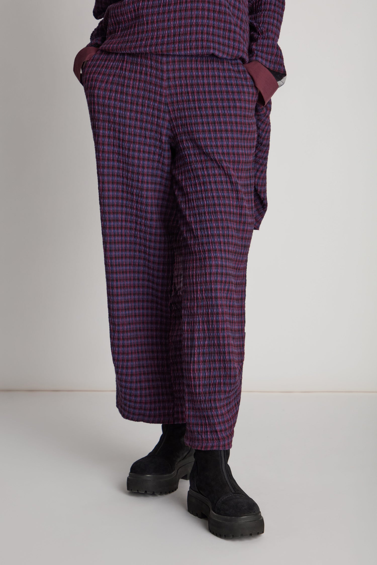 Individual dressed in the Check Trouser—featuring a purple and black checkered pattern—and black boots, with hands tucked in pockets. Upper torso is not visible.