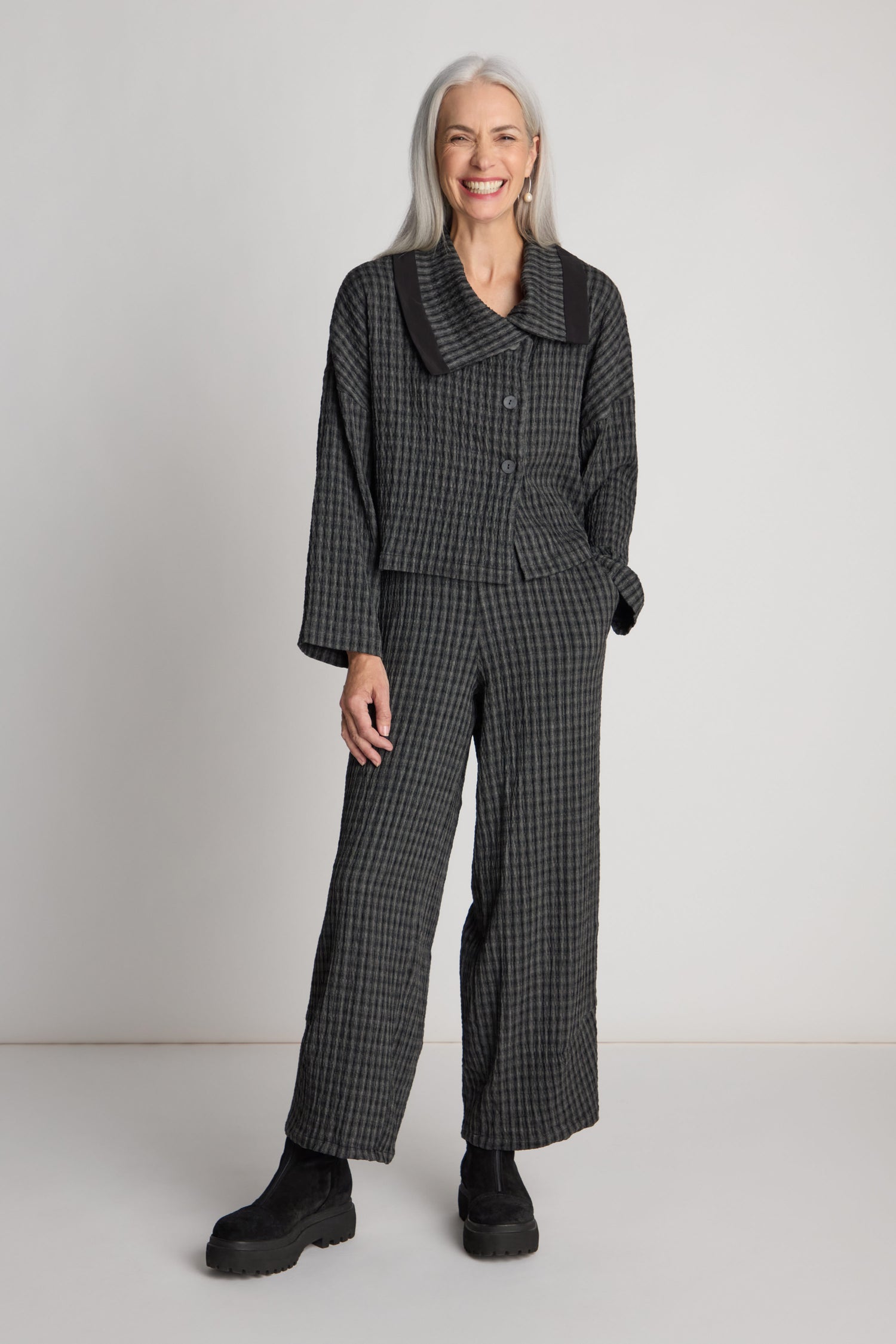 A person with long gray hair stands smiling, wearing the Check Shawl Collar Jacket and wide-legged pants in gray checkered gauze fabric, black ankle boots, and hands in pockets.