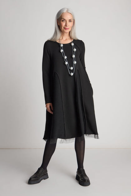 A woman with long gray hair is wearing a Godet Net Hem Jersey Dress with an A-line silhouette, along with black tights, black shoes, and a silver necklace, standing against a plain white background.