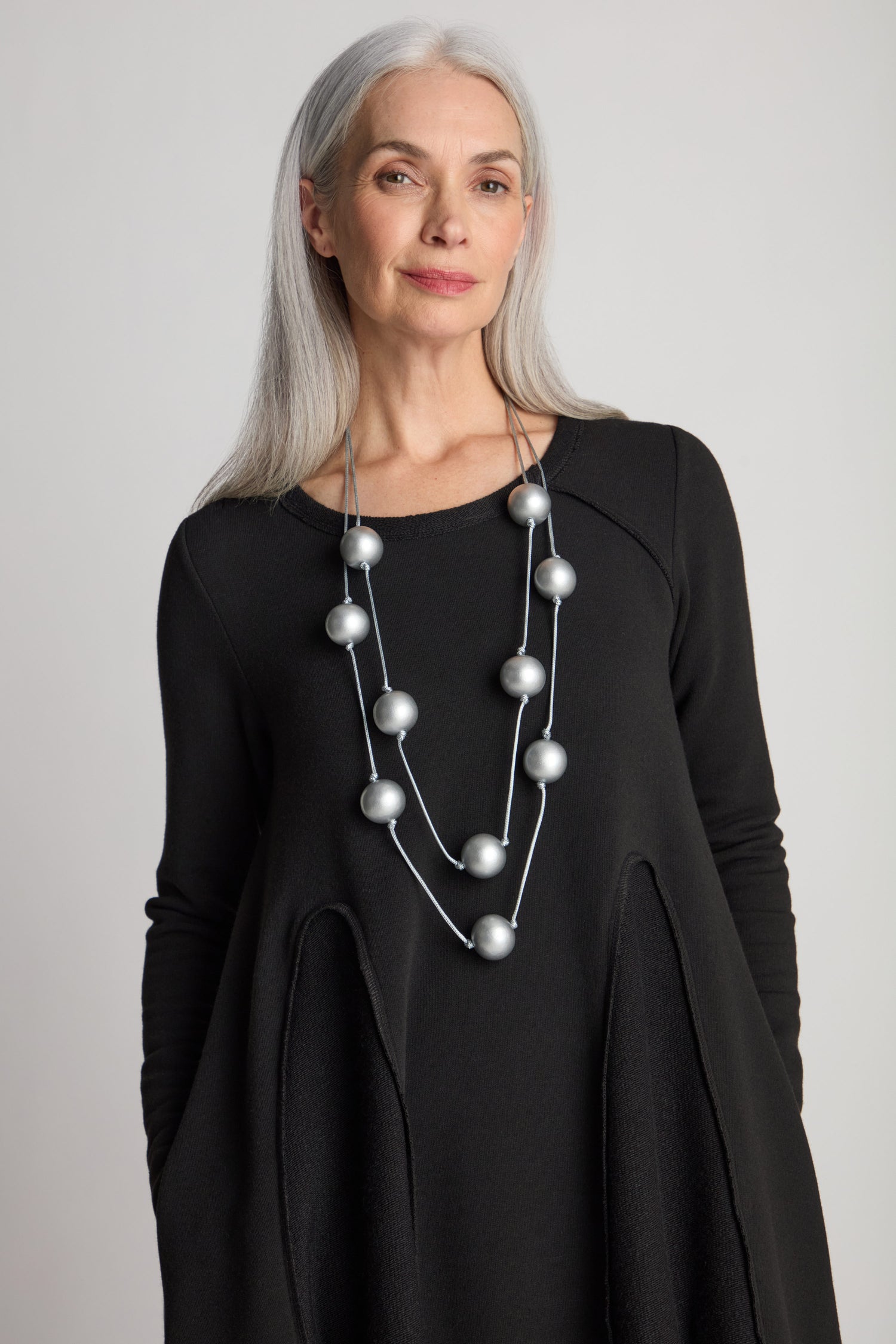 An older woman with long gray hair, wearing a black Godet Net Hem Jersey Dress featuring an A-line silhouette and a large silver bead necklace, poses against a plain background.