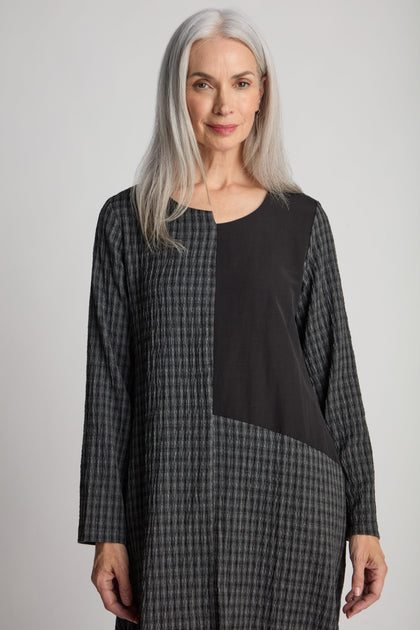 An older woman with long gray hair is standing and looking at the camera. She is wearing a Patched Check Dress with long sleeves, featuring a plaid pattern, a solid black panel, and an elegant asymmetric hem.