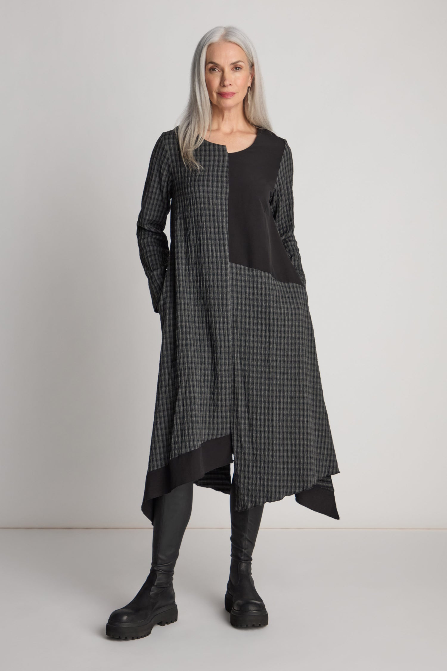 A woman with long gray hair stands against a plain background, wearing the Patched Check Dress featuring a loose-fitting asymmetric hem and contrasting patchwork design, paired with black leggings and black boots.