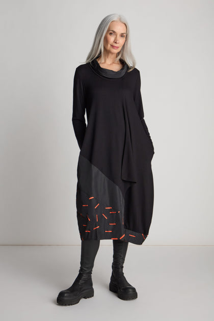 A woman with long gray hair is wearing a bold Cut Out Cowl Neck Dress, featuring orange stitching details and matched with black boots, as she stands against a plain white background.