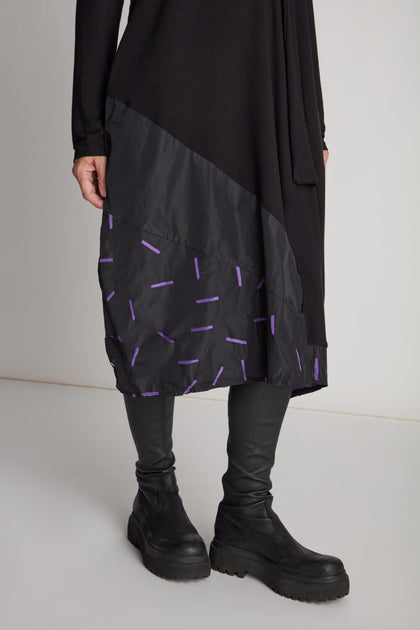 A person wearing a Cut Out Cowl Neck Dress with a purple pattern on the lower part, paired with black leggings and black boots, making it a statement piece.