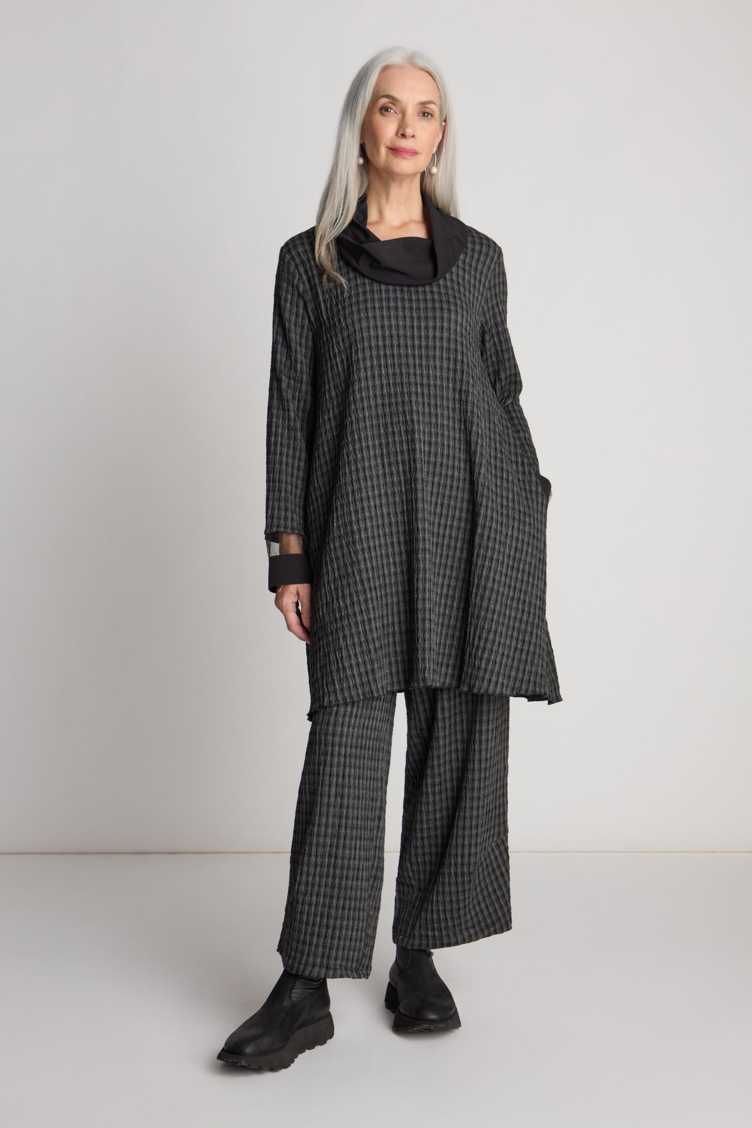 A person with long gray hair stands against a plain background, showcasing a checkered grey Luukaa Satin Cowl Check Tunic paired with wide-leg pants and black shoes—a testament to sustainable fashion.