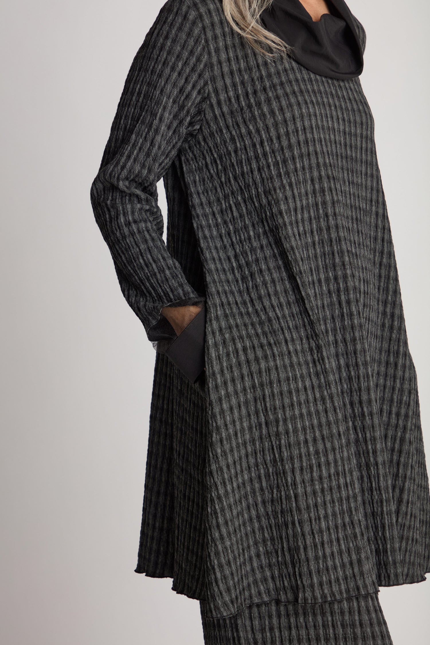 A person wearing Luukaa's sustainable fashion piece, the Satin Cowl Check Tunic, featuring long sleeves with a black and grey checkered design and a black collar, stands with their hands in their pockets.