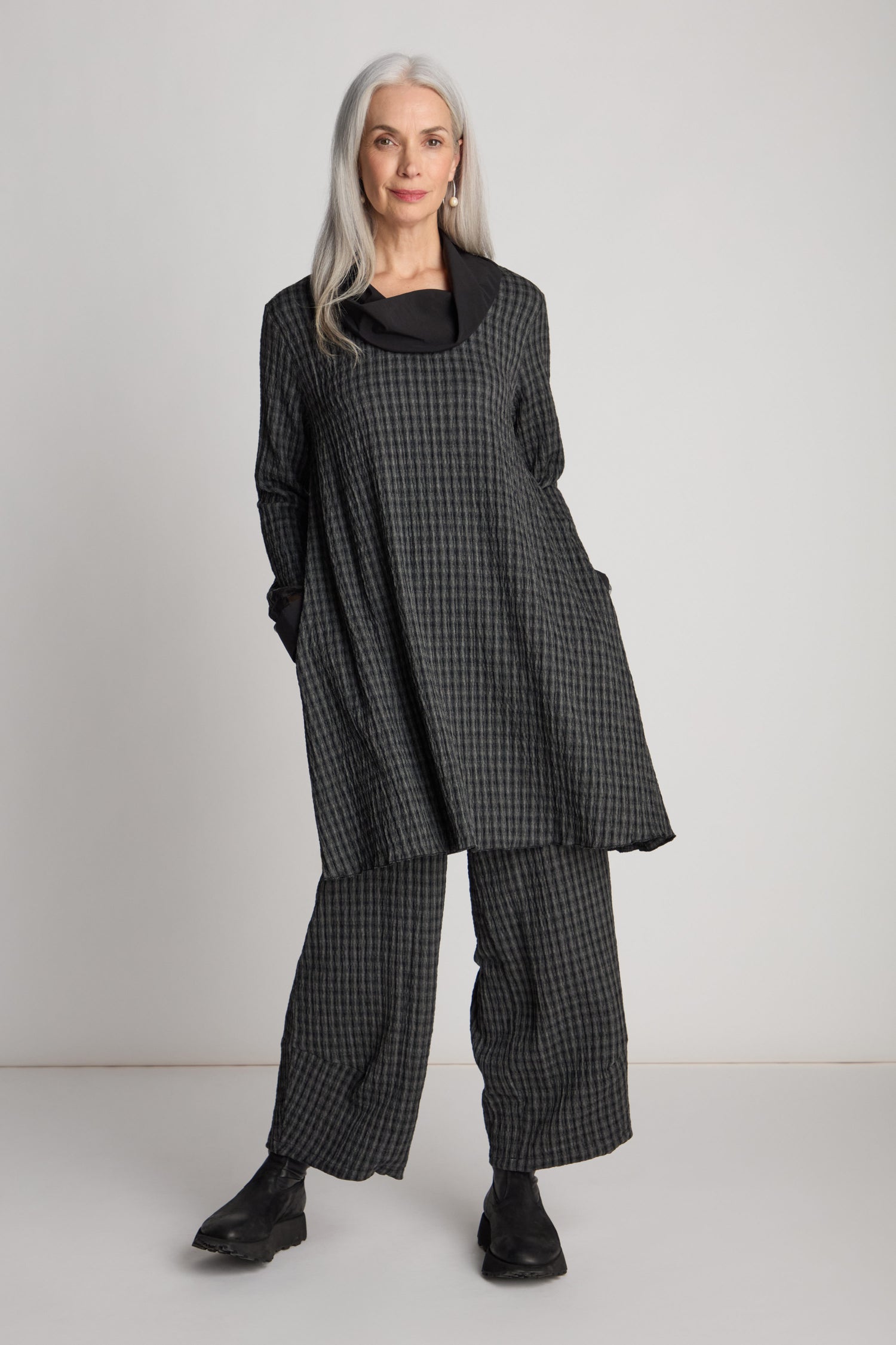 A woman with long gray hair is wearing the Satin Cowl Check Tunic from Luukaa, paired perfectly with matching wide-leg pants. She stands with her hands in her pockets against a plain background, effortlessly showcasing sustainable fashion.