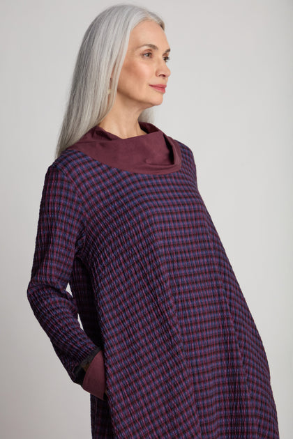 A woman with long gray hair stands in a side profile, wearing the Satin Cowl Check Tunic by Luukaa. She embodies sustainable fashion as she looks ahead with one hand in her pocket.