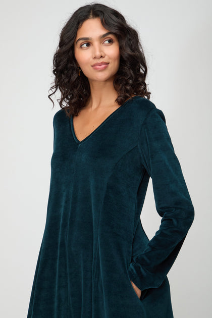 Woman with curly dark hair wears the V Neck Velvet Jersey Bubble Dress with long sleeves, standing against a plain light background and looking slightly to the side.