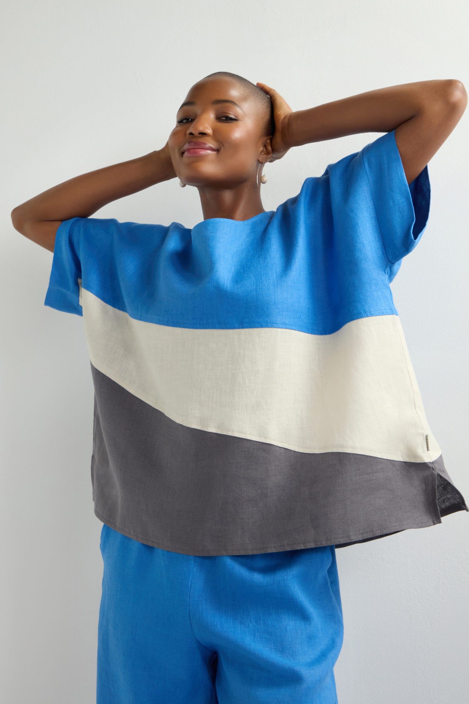 Wearing the Colour Block Top in blue, white, and gray with matching blue pants, they stand confidently with hands behind their head and a slight smile, exemplifying contemporary fashion.