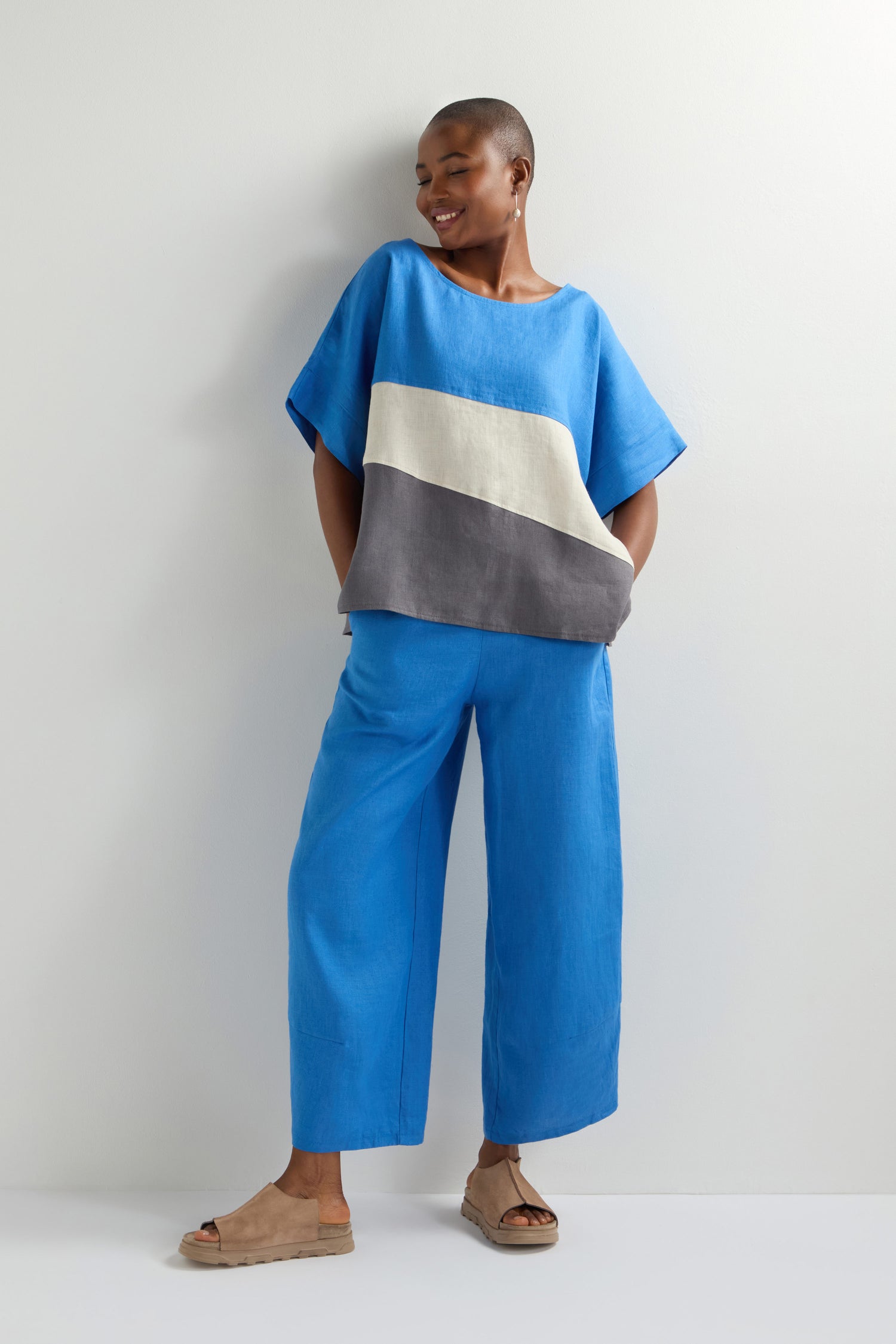 A person dressed in contemporary fashion showcases the Colour Block Top in blue, standing against a plain backdrop. They smile subtly while posing confidently with one hand in their pocket, turning the ensemble into a statement piece.