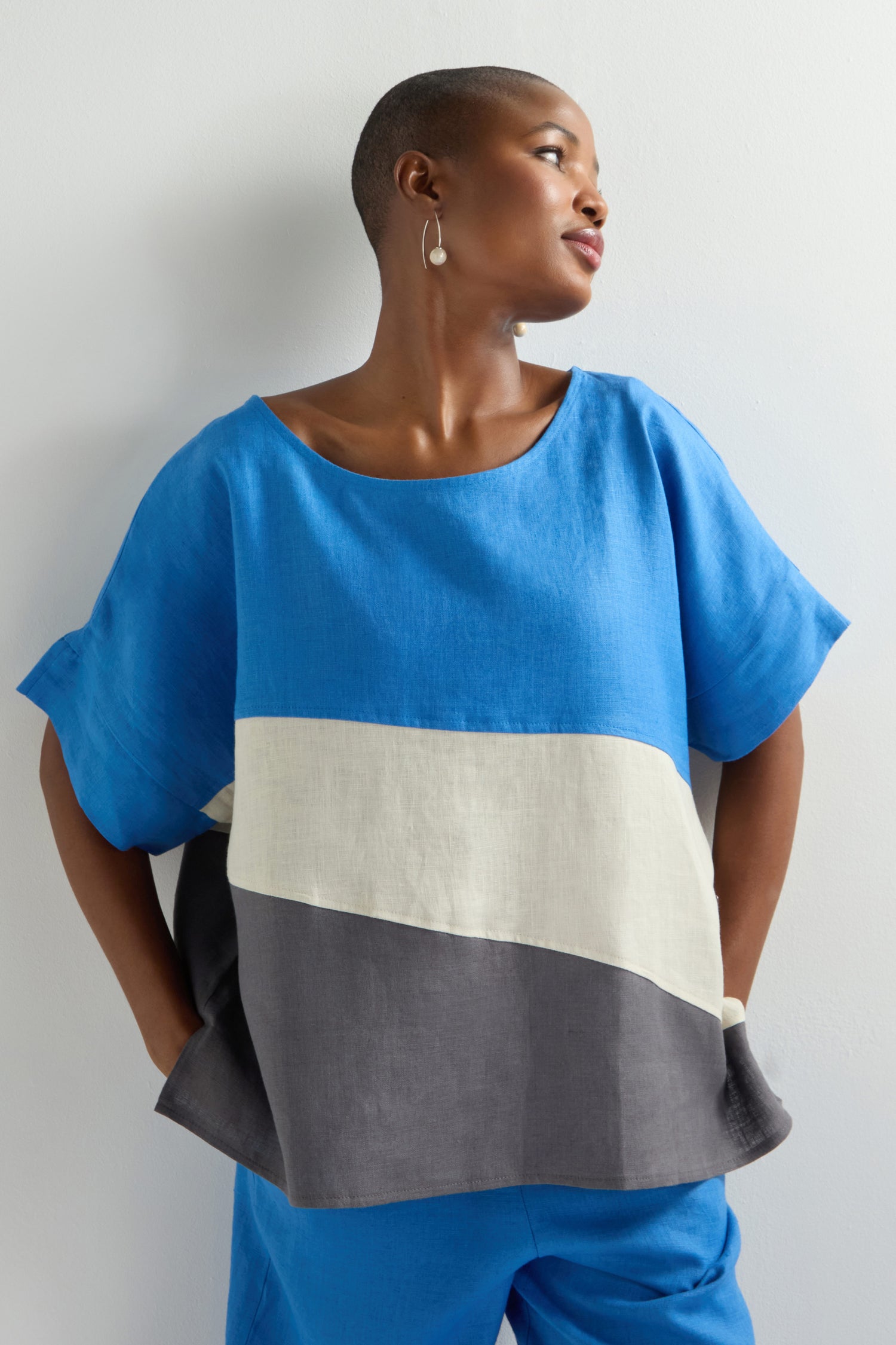 A person clad in the "Colour Block Top" in blue, white, and gray pairs it with blue pants against a plain backdrop, epitomizing contemporary fashion. This standout piece elegantly encapsulates modern style's essence.