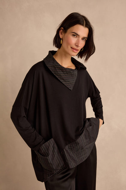 A woman with short dark hair sits on a wooden chair against a neutral background, wearing the Stripe Jersey Taffeta Tunic in loose brown and black stripes.