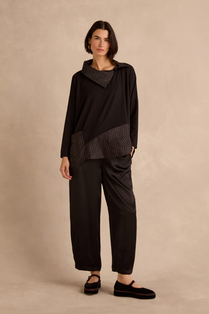A woman with short dark hair and a neutral expression stands against a beige background wearing the Stripe Jersey Taffeta Tunic featuring black stripes, paired with black pants with elastic cuffs and black shoes.