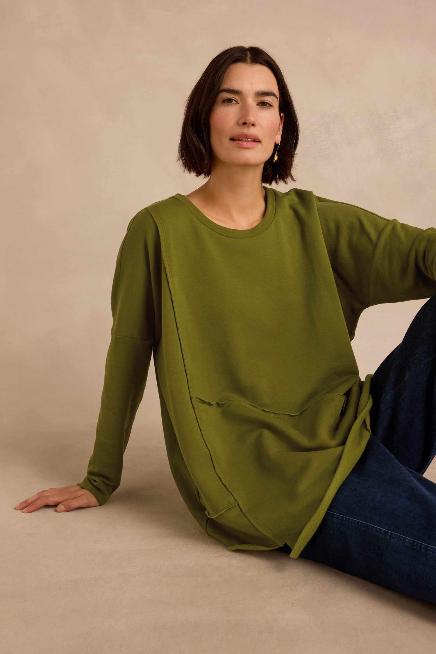 A woman sits on the floor against a neutral background, wearing dark jeans and the Seam Detail Pocket Top by Luukaa—a loose-fitting green shirt crafted from cotton lyocell stretch jersey.