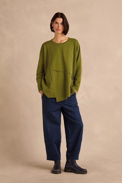 A person stands against a beige background wearing a loose green Seam Detail Pocket Top, dark blue wide-leg jeans, and black boots, with hands in pockets and short dark hair.
