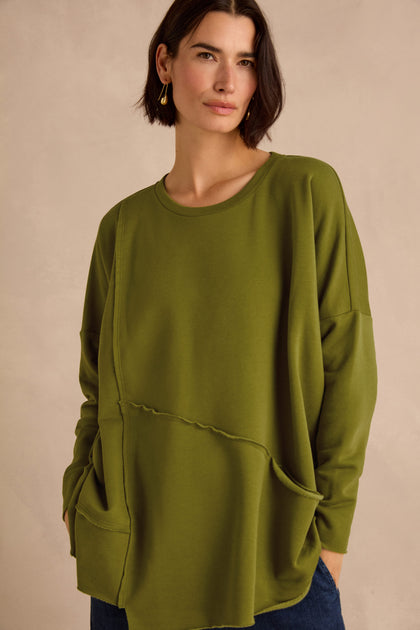 A woman stands against a neutral background, wearing the Seam Detail Pocket Top in an oversized olive green silhouette, crafted from cotton lyocell stretch jersey and featuring two large front pockets.