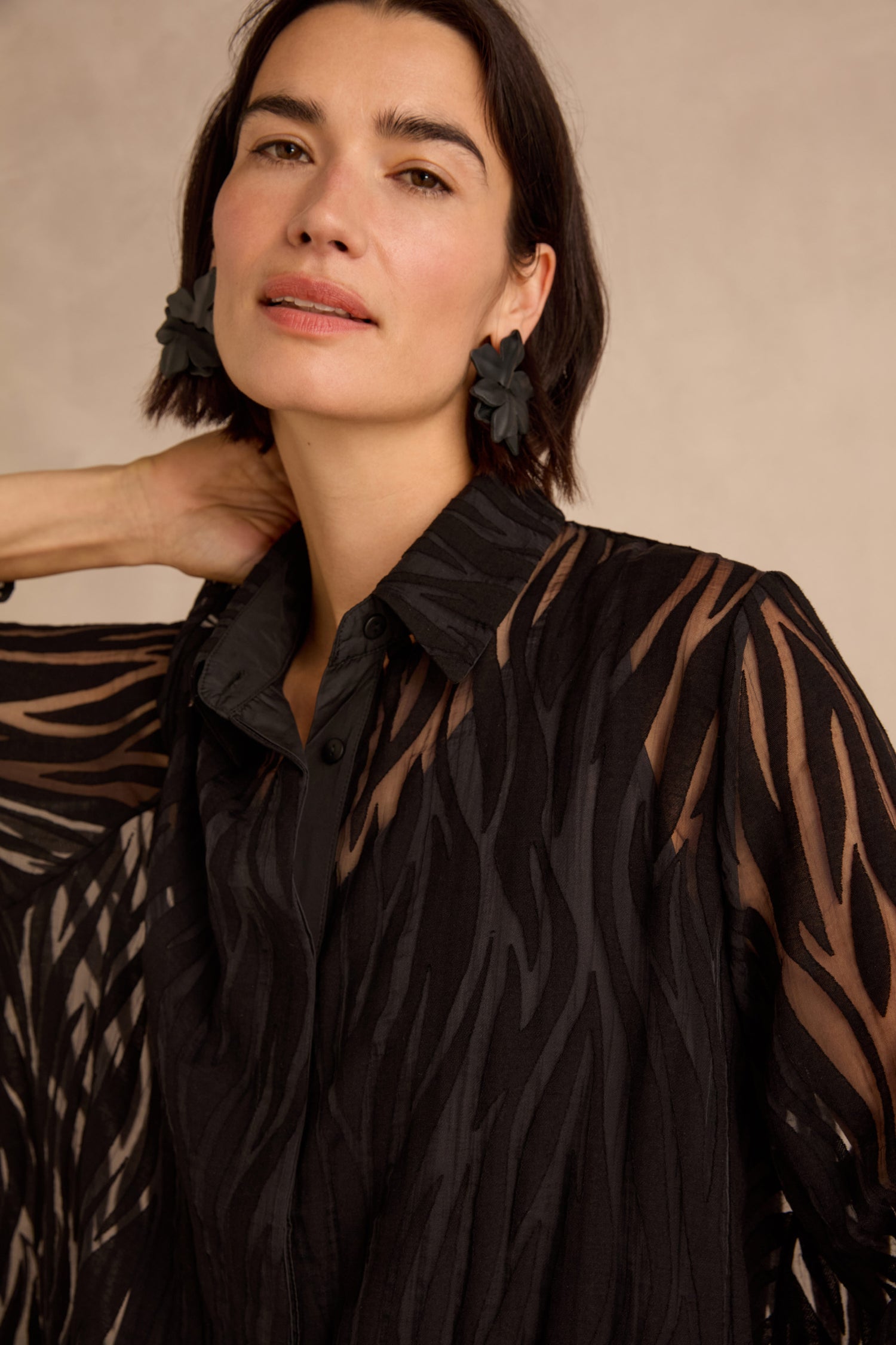 Woman with short dark hair wearing the Wave Cut Out Shirt—a sheer, tunic-length, black patterned blouse crafted from a viscose blend—paired with flower-shaped black earrings, standing against a neutral background.