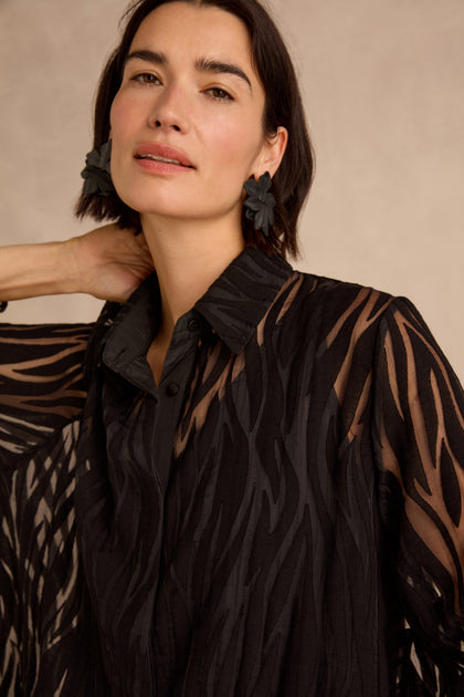 Woman with short dark hair wearing the Wave Cut Out Shirt—a sheer, tunic-length, black patterned blouse crafted from a viscose blend—paired with flower-shaped black earrings, standing against a neutral background.