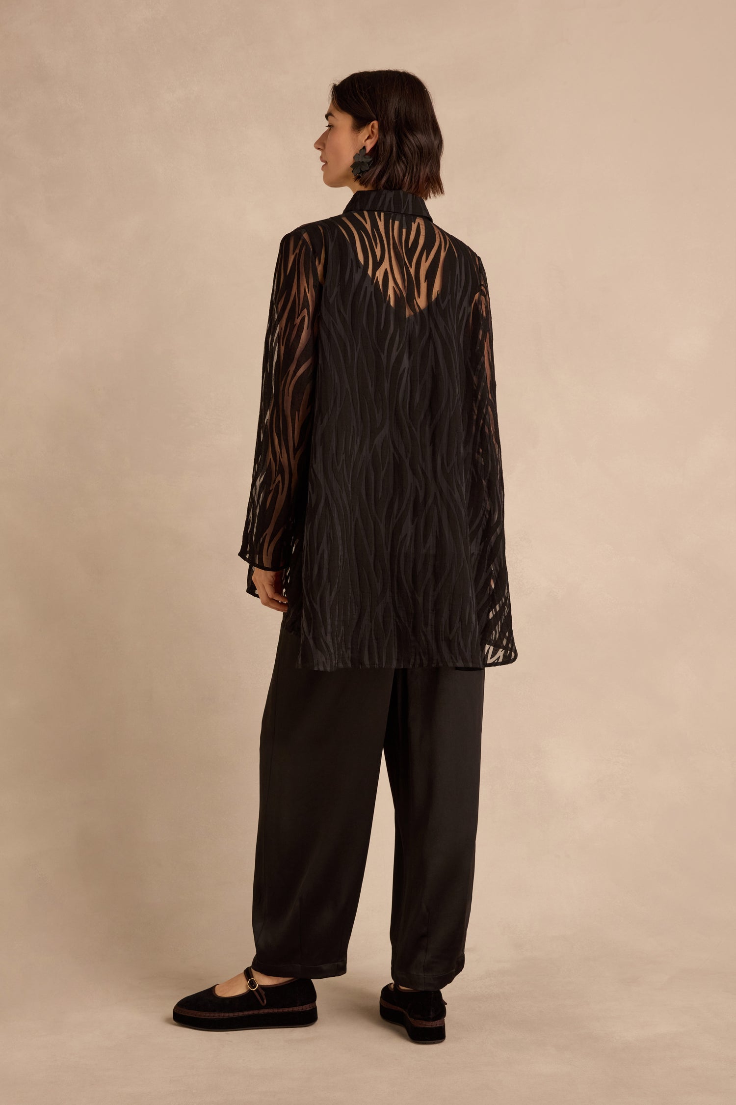 A woman stands against a neutral background, facing away from the camera, wearing black loose pants, black shoes, and the Wave Cut Out Shirt—a sheer viscose-blend tunic-length design—layered over a black shirt.